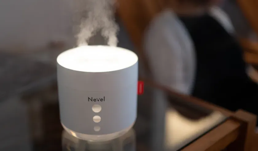 白い加湿器から蒸気が出ている様子を捉えた写真です。加湿器はテーブルの上に置かれており、背景にはぼやけた人物がいます。