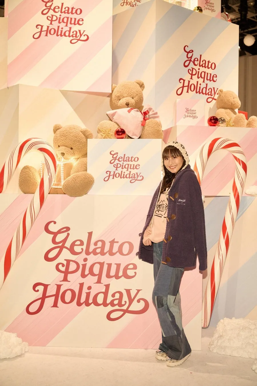 女性が「Gelato Pique Holiday」と書かれた背景の前でポーズをとっています。背景にはテディベア、キャンディケーン、プレゼントが飾られています。