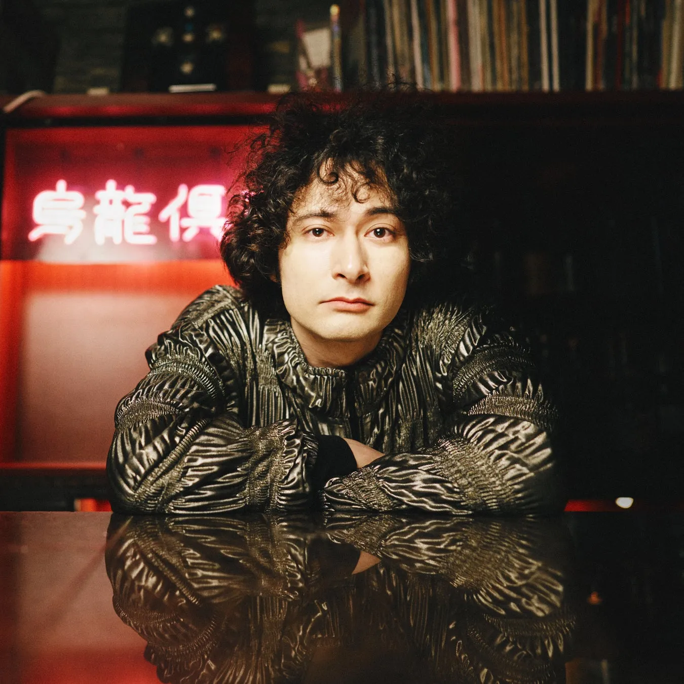 テーブルに腕を組み、カメラ目線で座っている男性のポートレート。背景にはネオンサインと本棚がある。