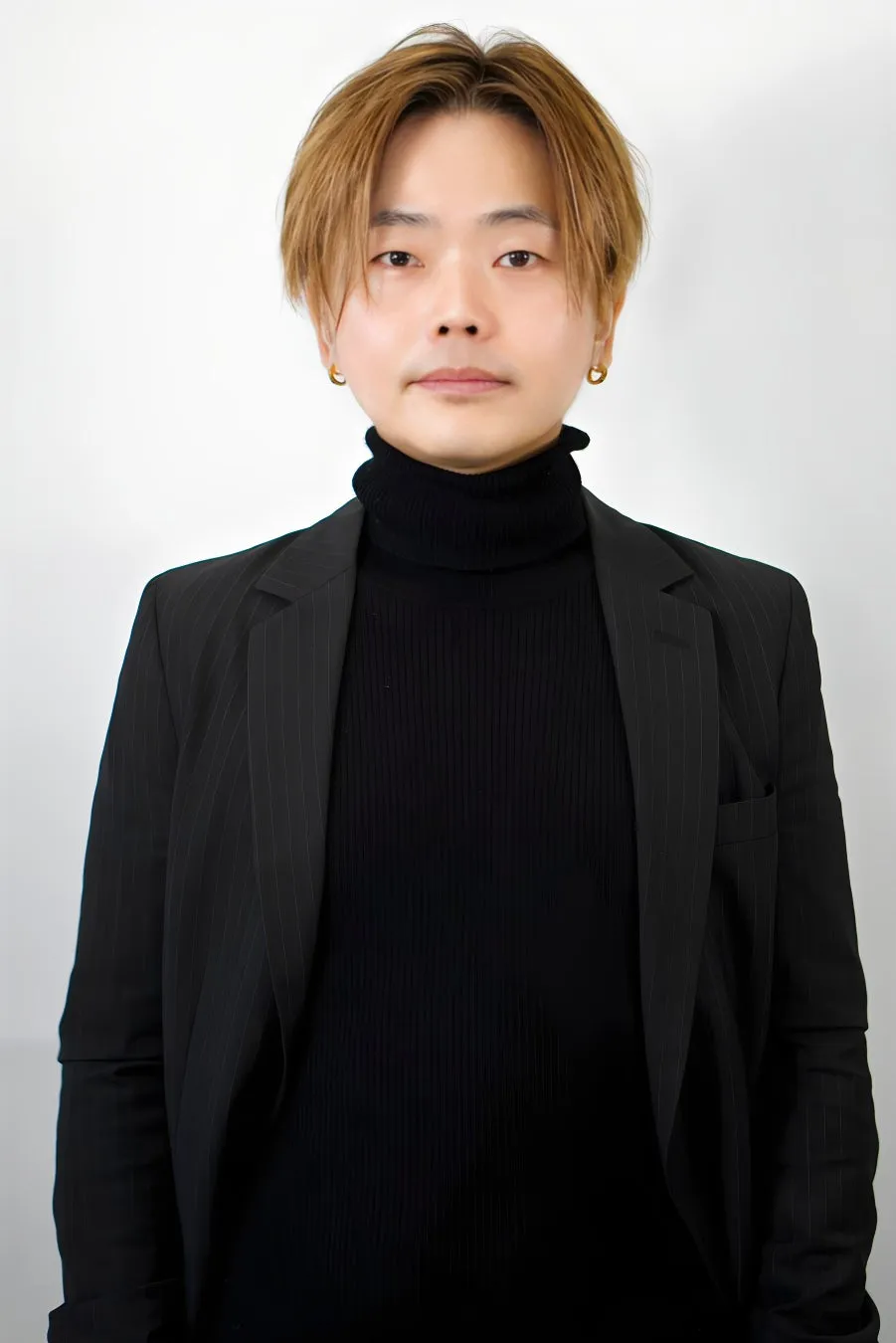 男性が正面を向き、黒いタートルネックセーターと黒いストライプのジャケットを着て立っています。背景は白です。