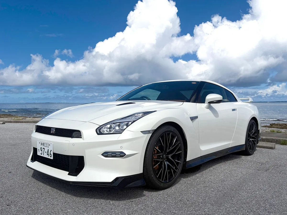 白い日産GT-Rが海辺の道に駐車されている写真です。空には雲が広がり、車と海のコントラストが美しいです。