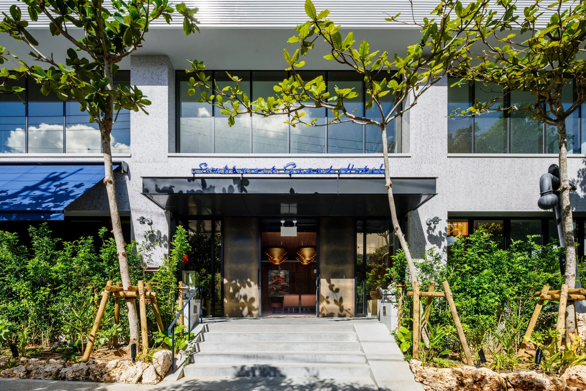 ホテルの正面玄関の写真です。ホテルの名前が看板に表示され、入り口には階段があります。建物は石造りで、緑豊かな木々が植えられています。