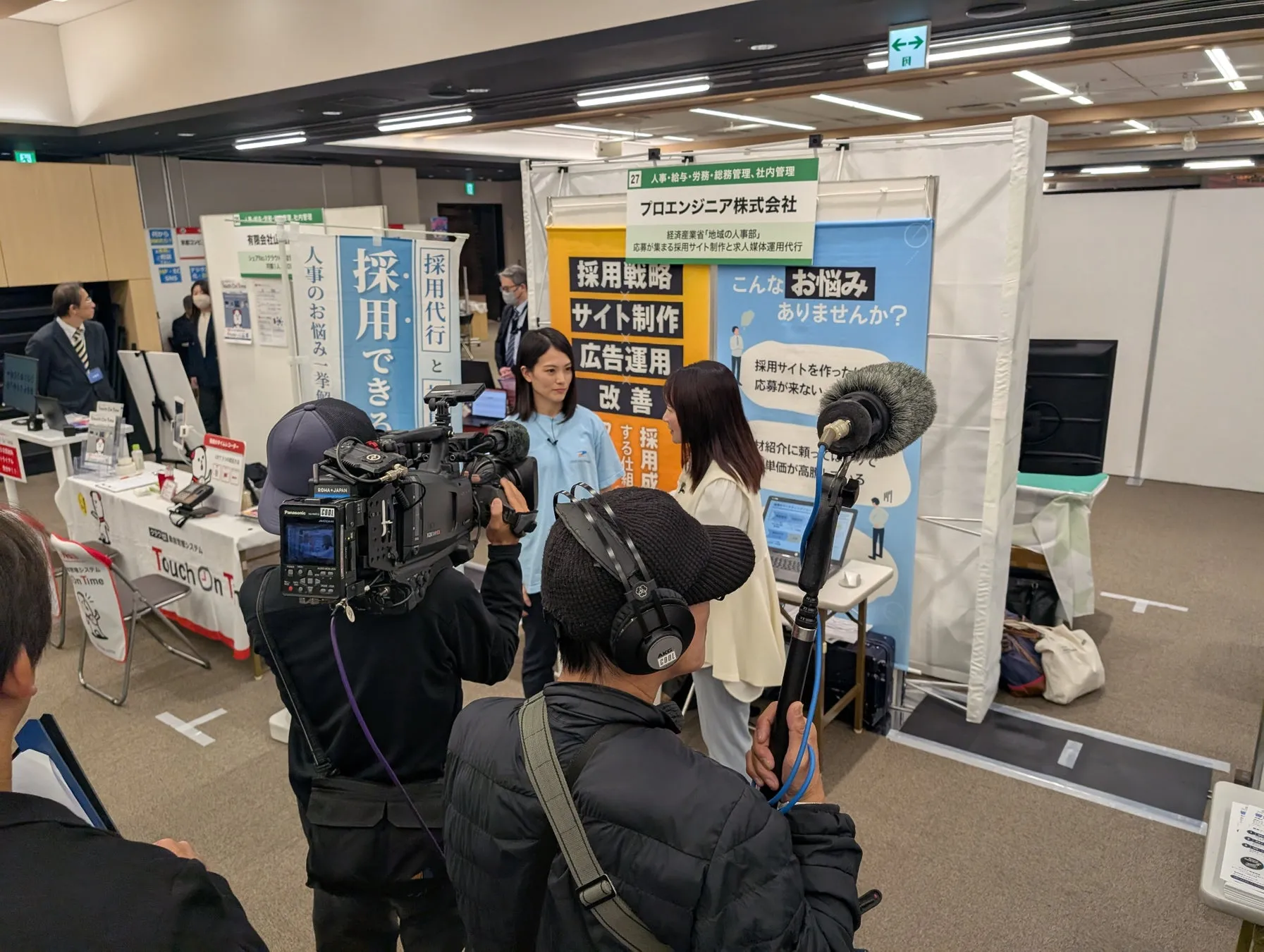 イベント会場で、プロエンジニア株式会社のブースで取材が行われている様子。採用戦略、サイト制作、広告運用、改善などのサービスが紹介されている。カメラマンや音声担当者、インタビューを受ける女性2名が写っている。