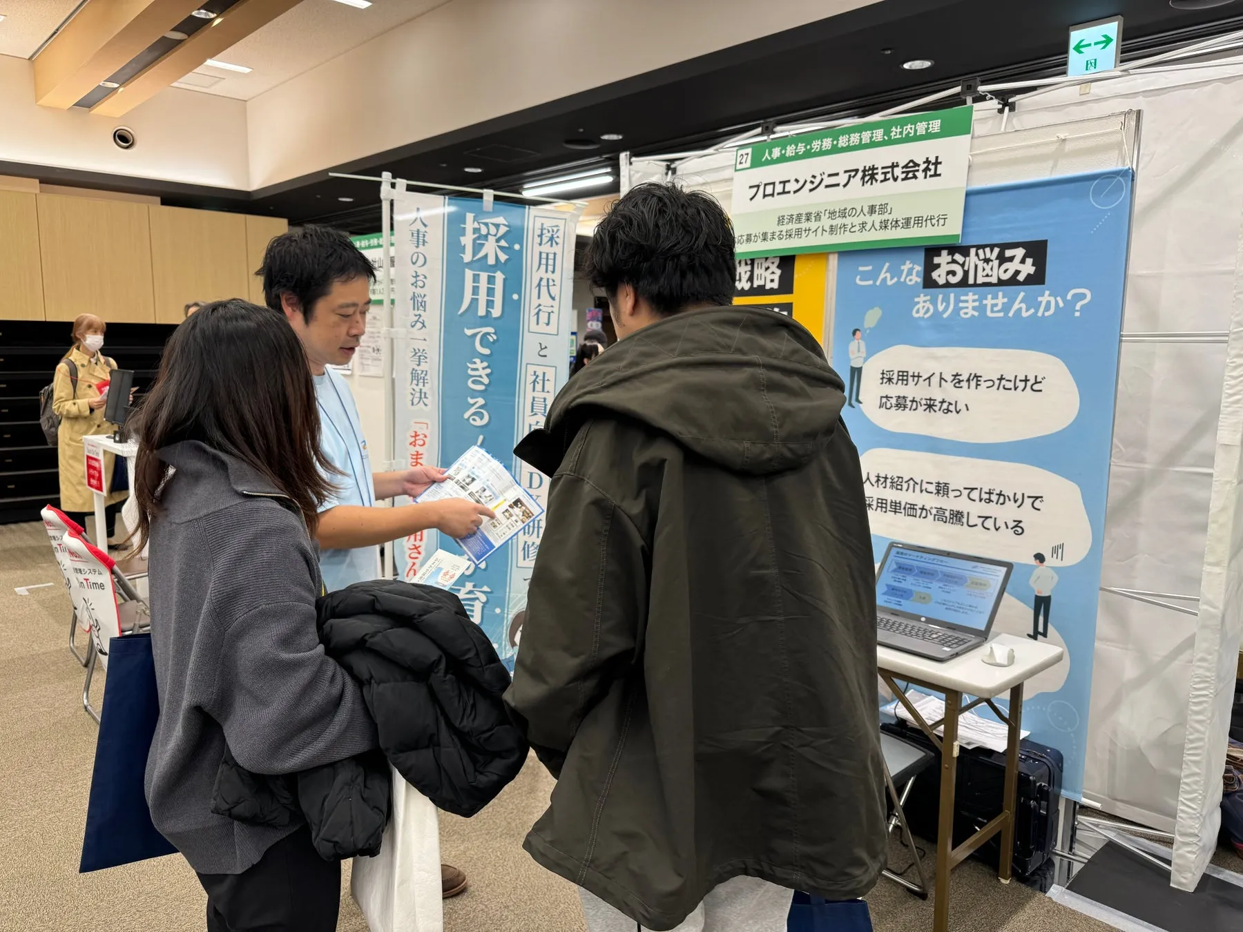展示会で、プロエンジニア株式会社の人事担当者が、来場者に採用に関する相談をしている様子。採用サイト制作や求人媒体運用代行、人事労務管理について相談できるようです。