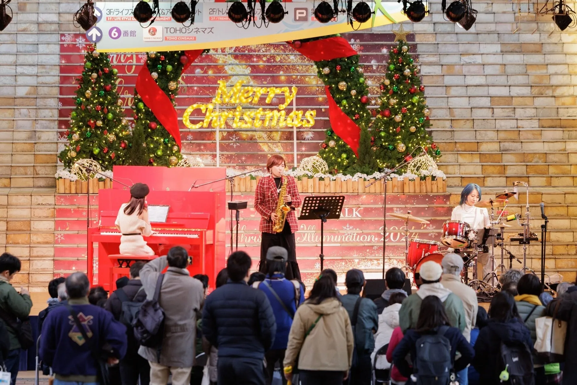 クリスマスイルミネーションの中で、サックスを演奏する女性と、ピアノ、ドラムの演奏者たちと観客の様子。