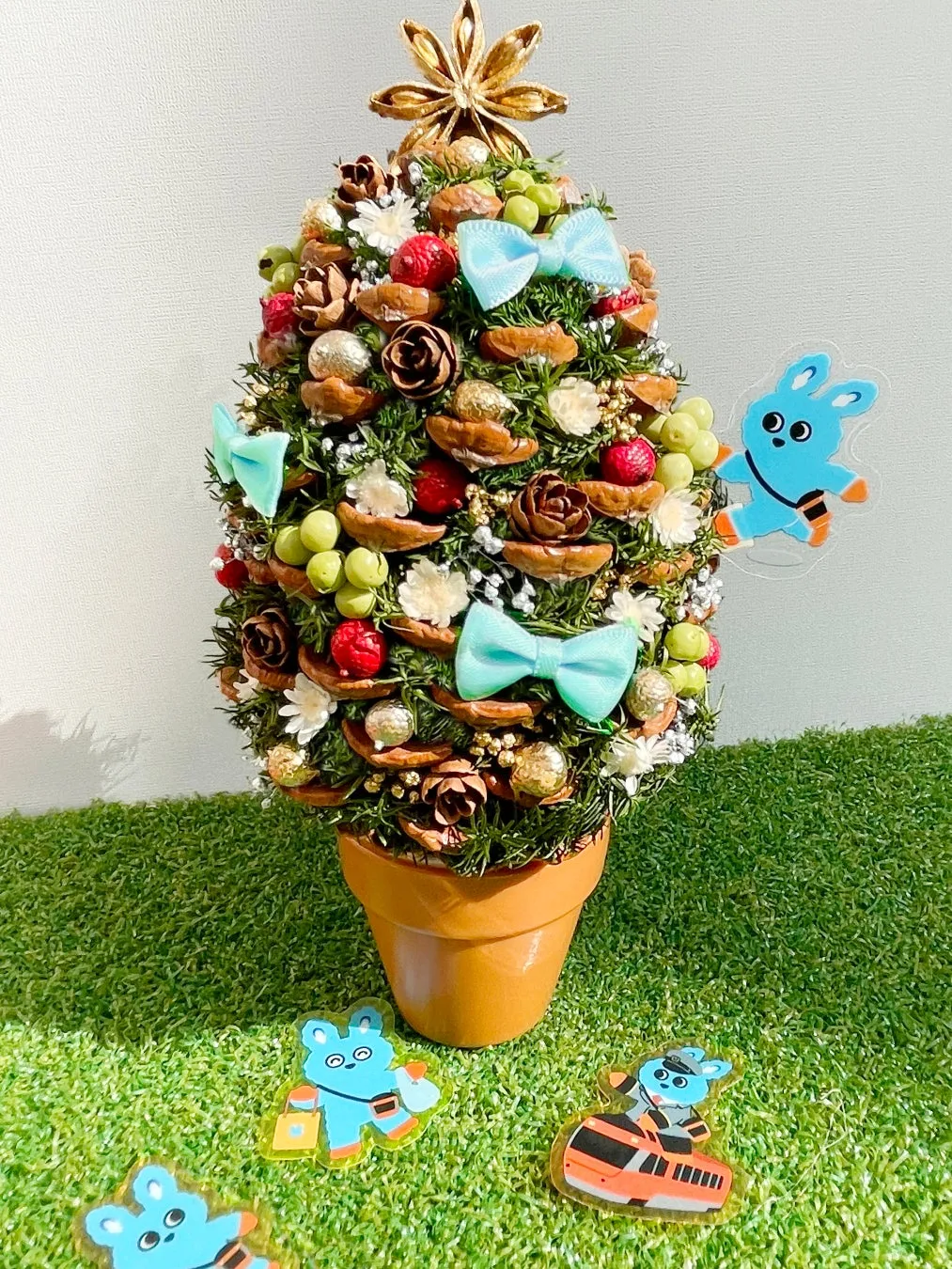 クリスマスツリーの形をした、松ぼっくり、リボン、装飾品、実で作られた装飾品。ツリーの周りには、青いキャラクターのステッカーがいくつか配置されている。