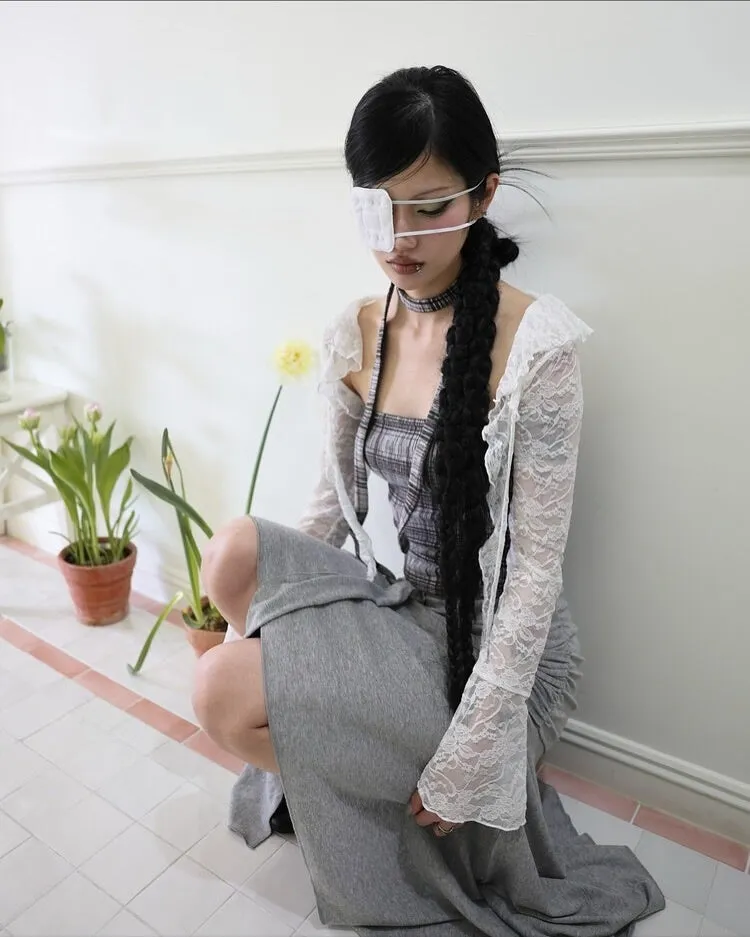 女性が片目に眼帯をし、長い黒髪を編み込み、床に座っている様子。白いレースのトップスとグレーのスカートを着用し、植物のそばにいる。