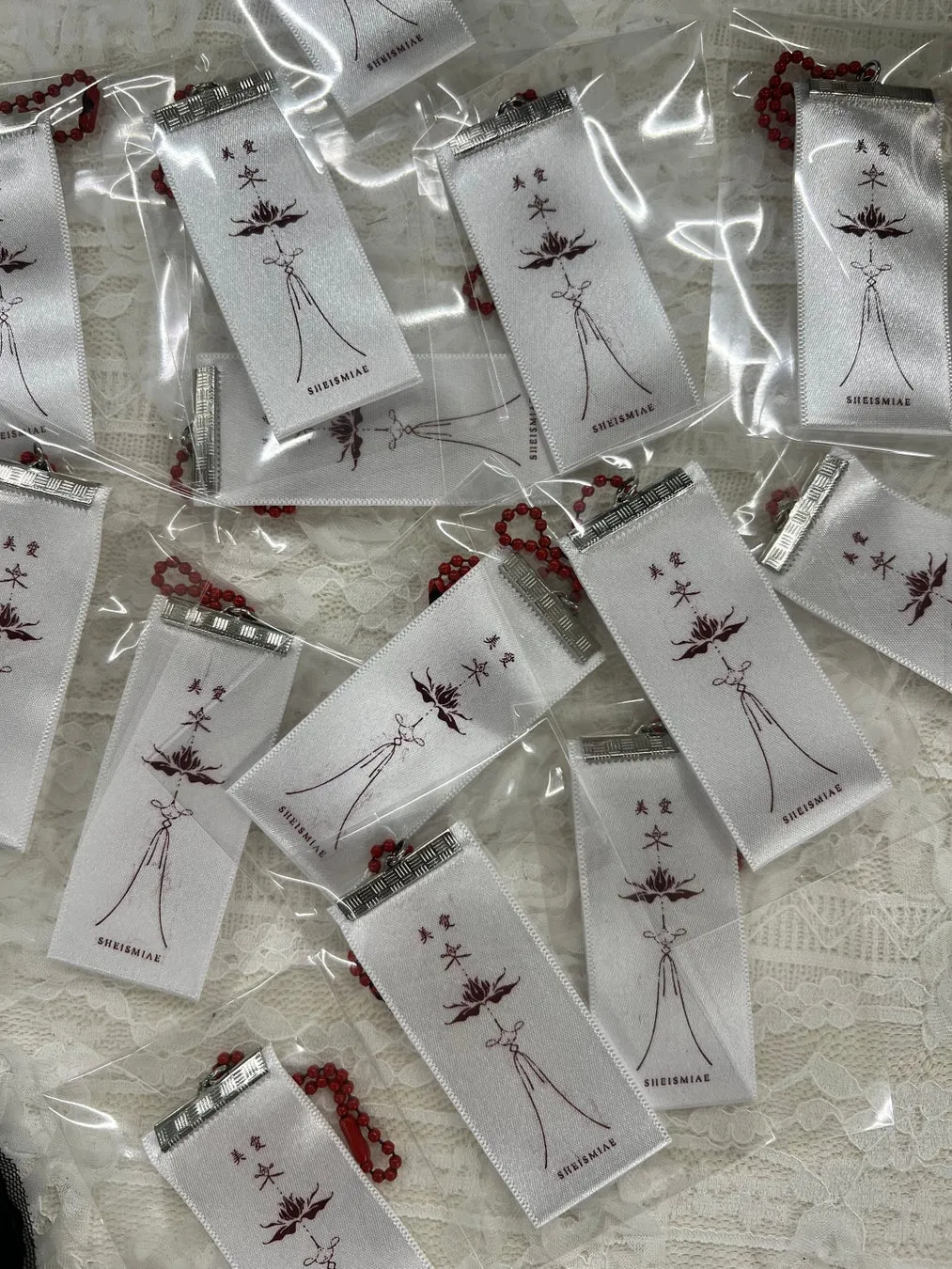 白いリボンに、赤い文字で「美愛」と蓮の花のようなイラストが描かれたアイテムが複数個写っている写真。リボンには「SHEISMIAE」の文字も記載されている。それぞれが透明な袋に入っており、赤いビーズのチェーンが付いている。