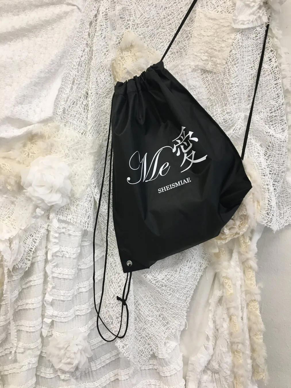 黒い巾着袋が白い布の上に置かれています。袋には「Me 愛 SHEISMIAE」と書かれています。