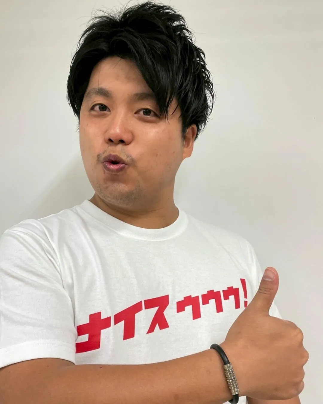 男性が親指を立てており、白いTシャツを着ており、Tシャツには赤い文字で「ナイスゥゥゥ!」と書かれています。