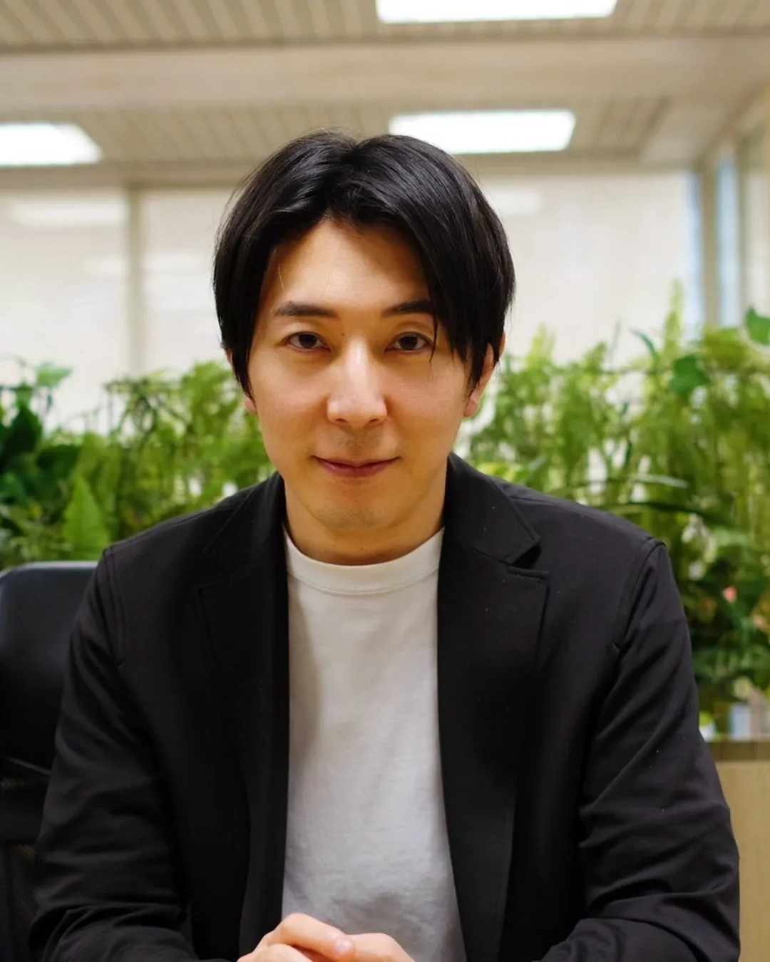 黒いジャケットを着た男性がカメラ目線で座っている写真です。背景には緑の植物と明るい照明が見えます。