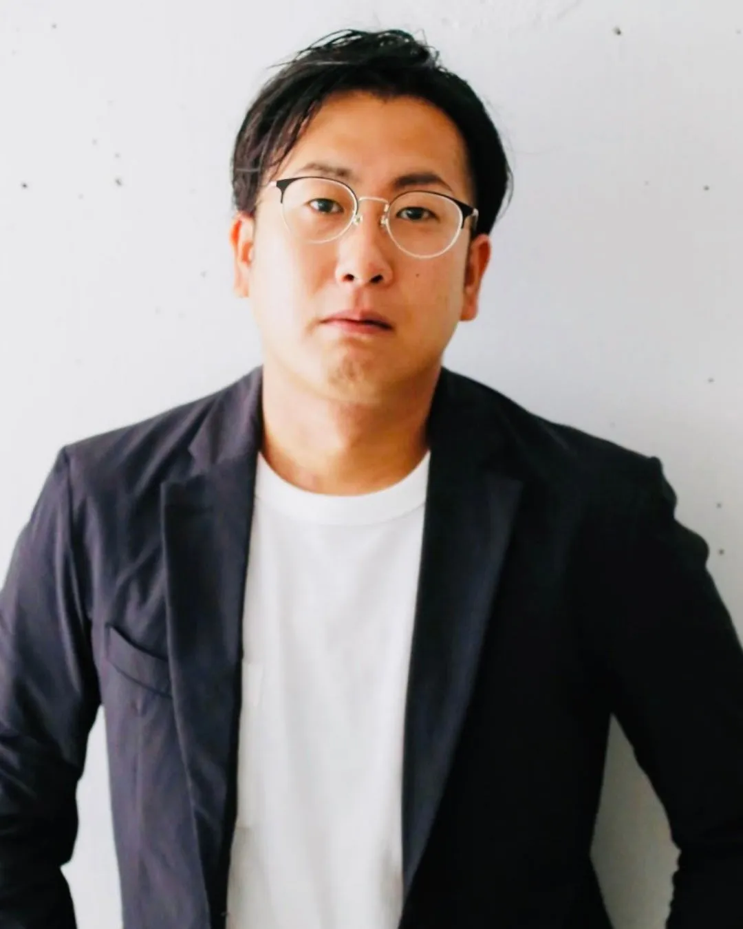 眼鏡をかけた男性がこちらを見ているポートレート写真です。彼は白いTシャツと黒いジャケットを着ています。