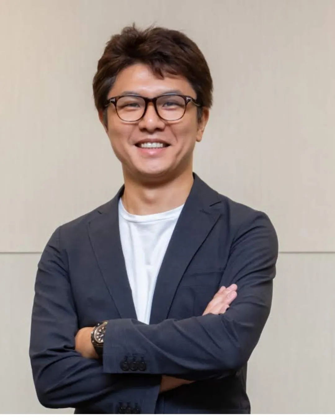 眼鏡をかけた男性が、腕を組んで笑顔で立っている様子。