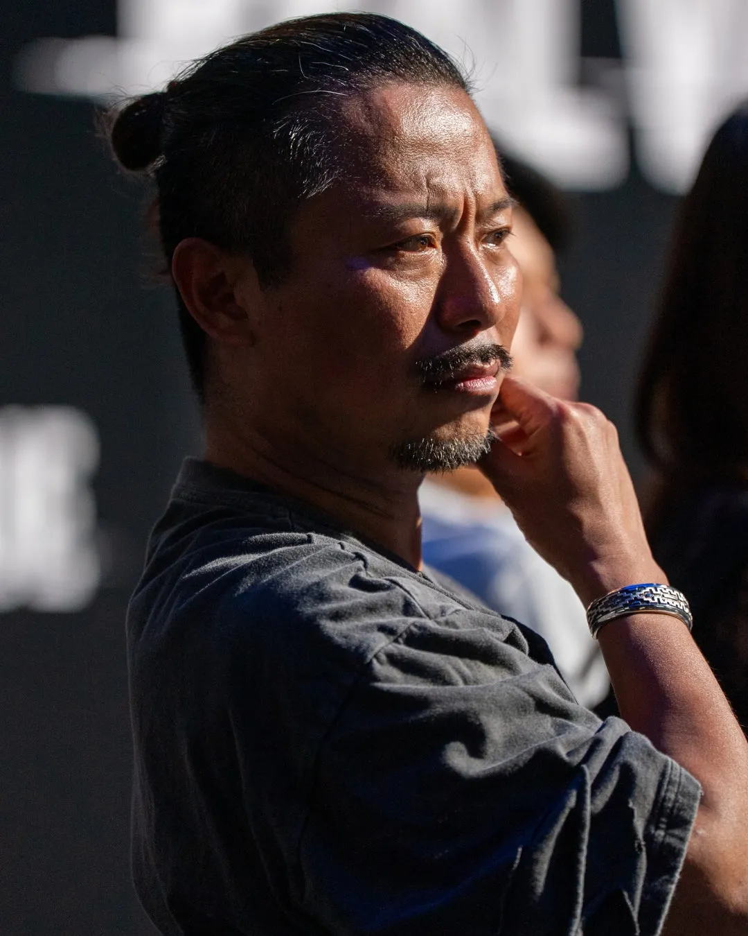 男性が腕を顎に当てて何かを考えている様子を捉えたポートレート。男性は少し上を見上げており、真剣な表情を浮かべている。背景には他の人物がぼやけて写っている。