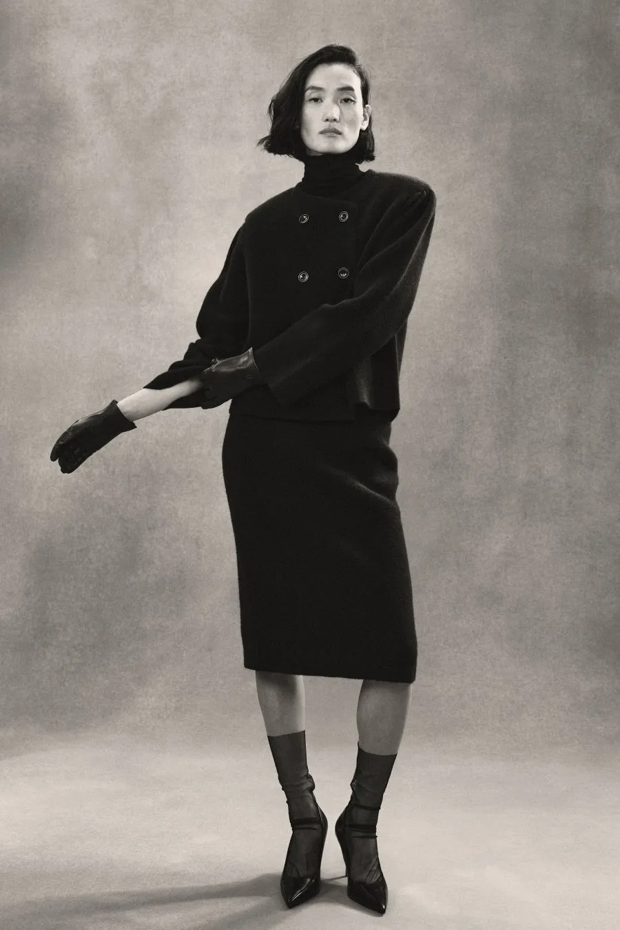 黒い服を着て、手袋をした女性の白黒写真。彼女はスカートとジャケットを着て、タートルネックのセーターを着ています。彼女は黒い靴を履いています。