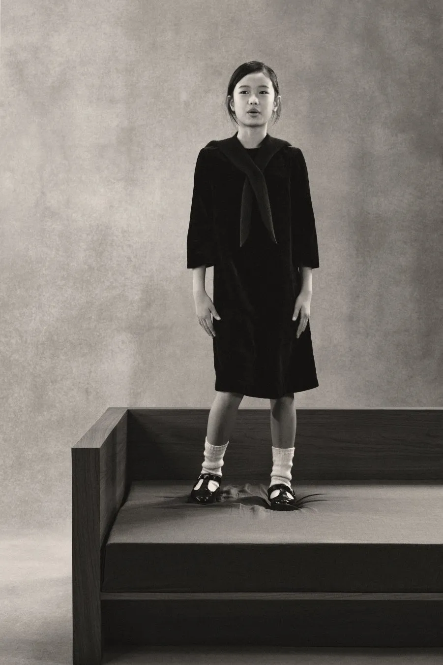 モノクロ写真。ベッドの上に立つ少女。少女は黒いワンピース、白い靴下、黒い靴を履いている。背景はグレー。