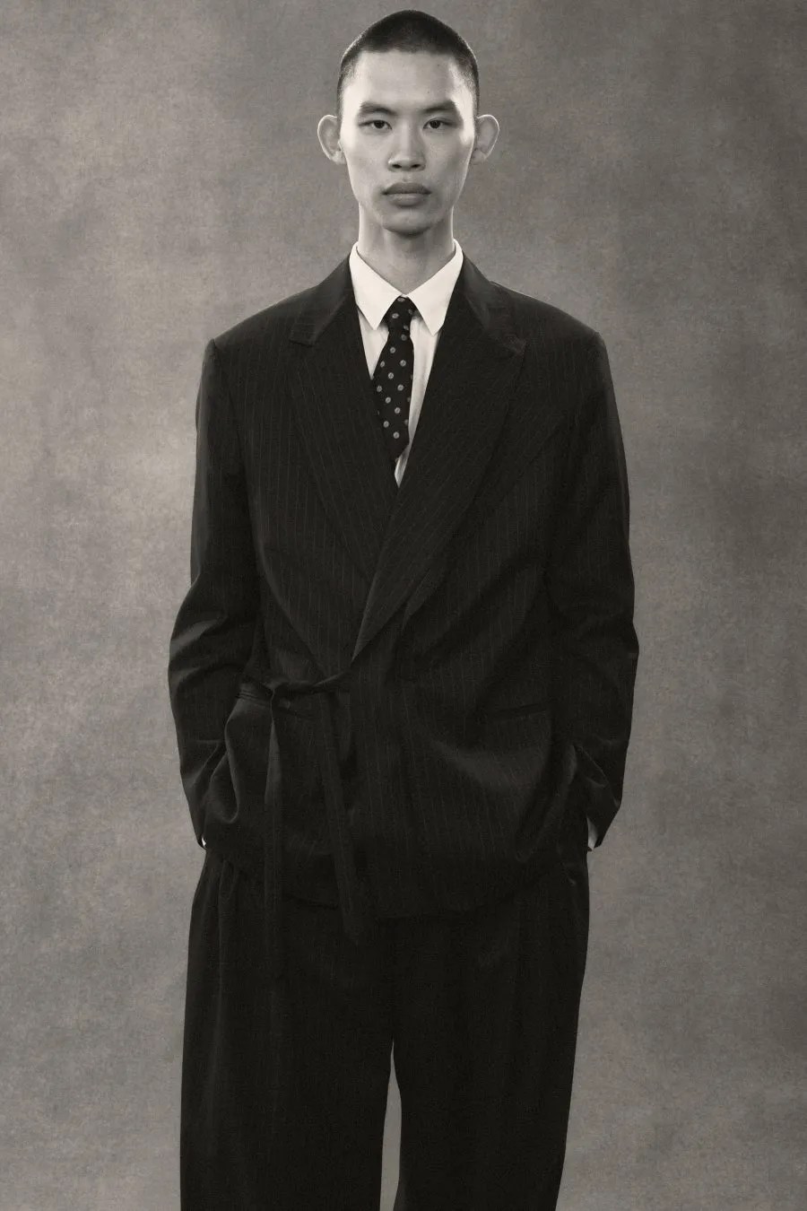 男性が黒いストライプのスーツを着て立っているモノクロ写真。彼は白いシャツとドット柄のネクタイを着用し、両手をポケットに入れている。