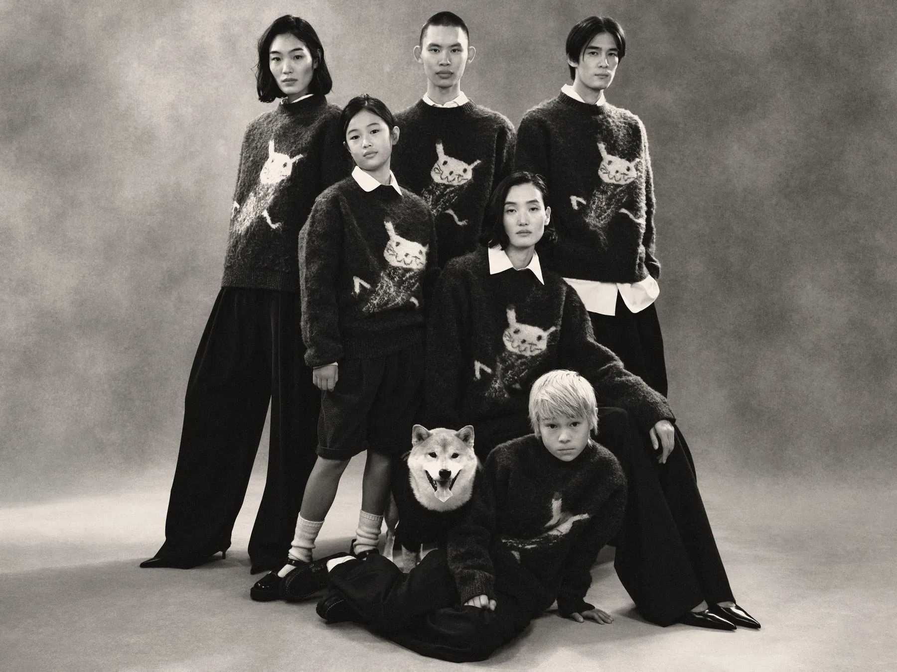 モノクロ写真には、ニットセーターを着た6人の人物と犬が写っています。犬はセーターを着ており、セーターには動物の顔のイラストが描かれています。人々はポーズをとってカメラを見ています。