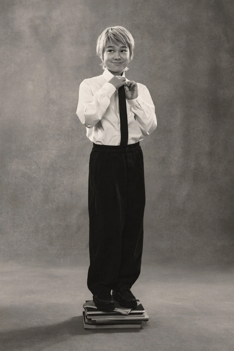 男の子が本の上に立ってネクタイを締めているモノクロ写真。