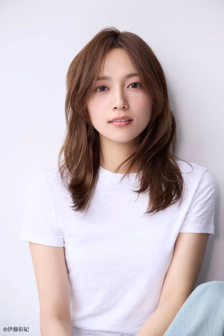白いTシャツを着た女性のポートレート。彼女はカメラに向かって微笑んでおり、長い茶色の髪が肩に流れている。背景は白。