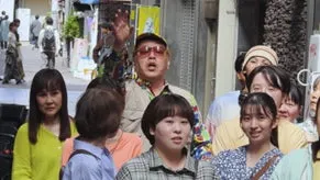 多くの人がいる中で、サングラスをかけた男性が手を上げて何かを話している様子。