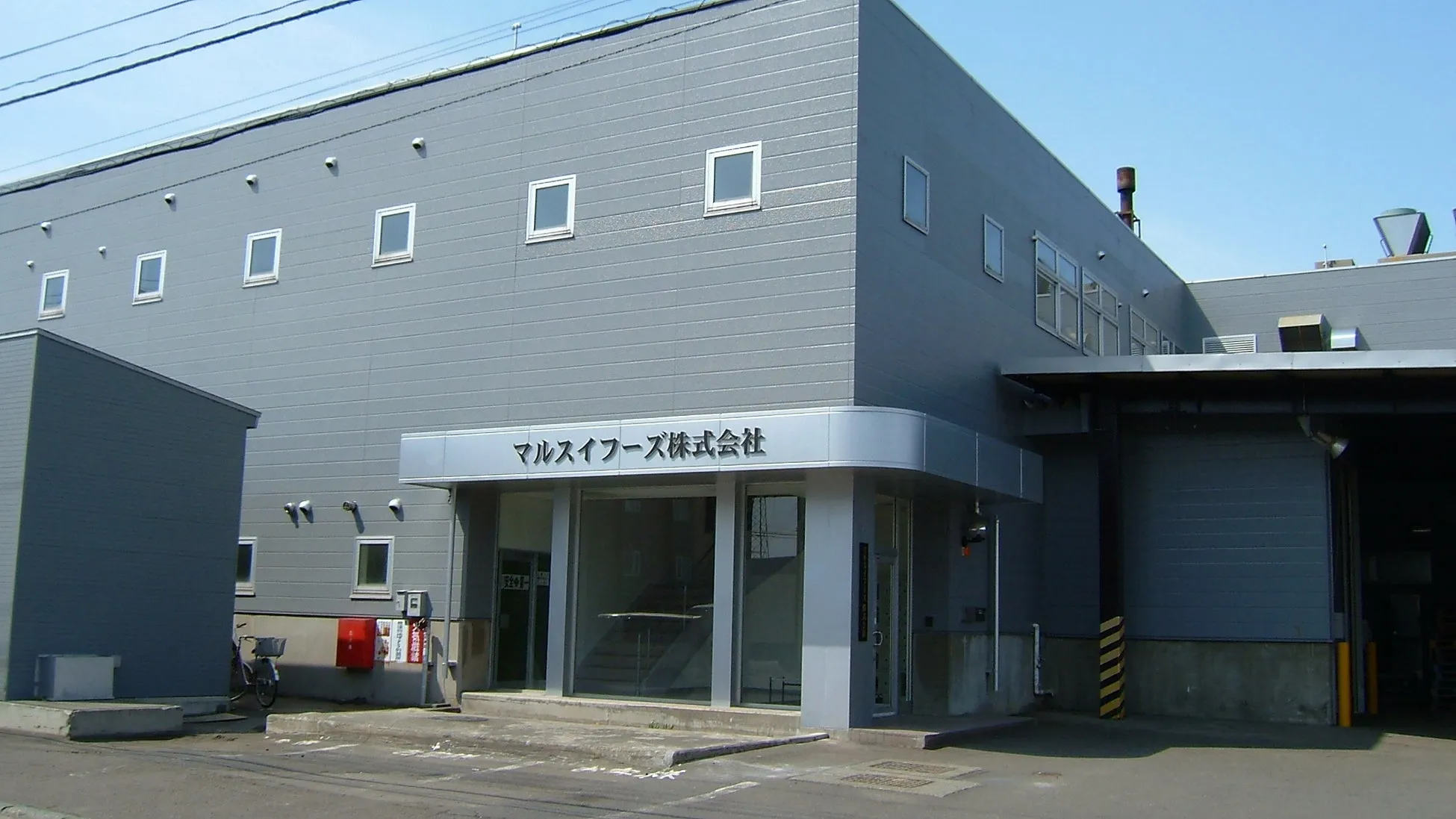 マルスイフーズ株式会社の建物の外観写真です。建物はグレーで、青空の下に建っています。