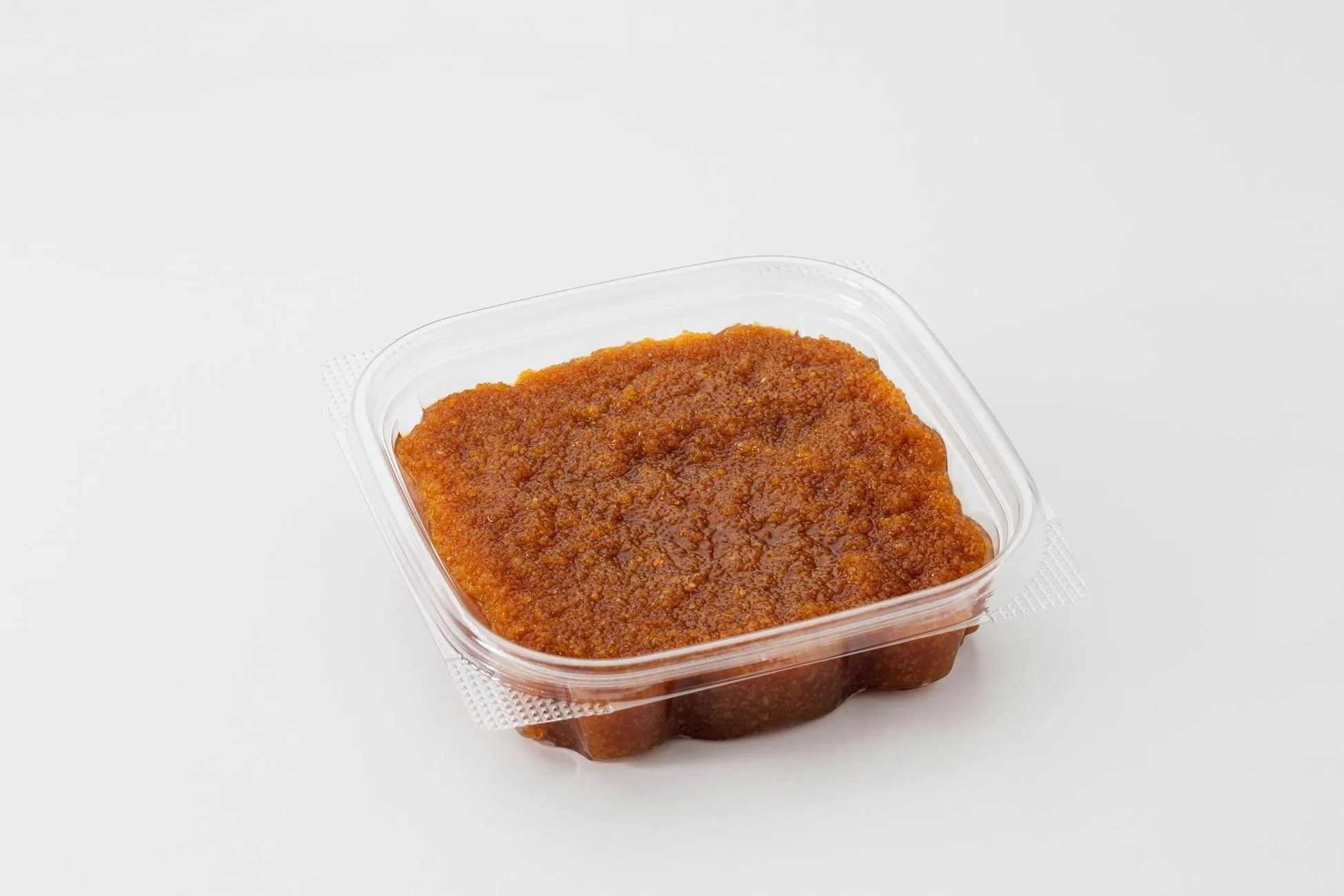 透明な容器に入った茶色の食品のクローズアップ写真です。食品は非常に細かく、粒状に見えます。容器は長方形で、白い背景に置かれています。