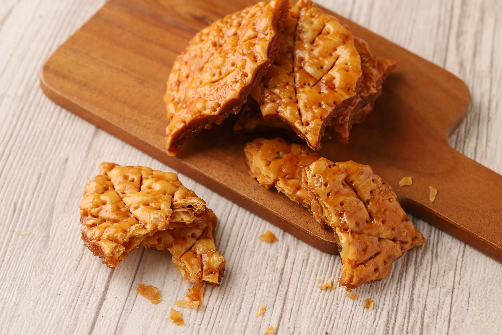 木製のまな板の上に、キャラメル色の焼き菓子がいくつか置かれています。焼き菓子は表面がツヤツヤしており、一部は割れて中身が見えています。背景は白木のテーブルで、焼き菓子の色合いが際立っています。