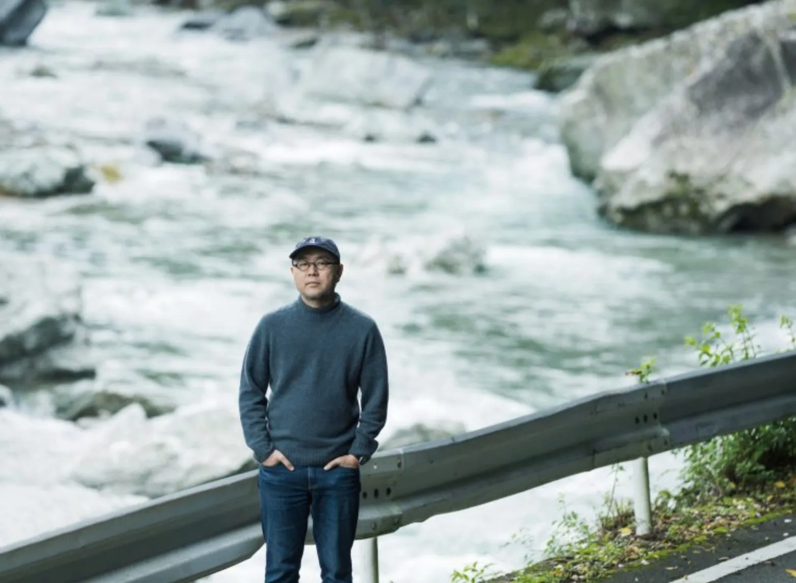 川のそばに立っている男性の画像です。彼は青いセーターとジーンズを着て、キャップをかぶっています。彼はカメラを見ています。背景には川と岩があります。