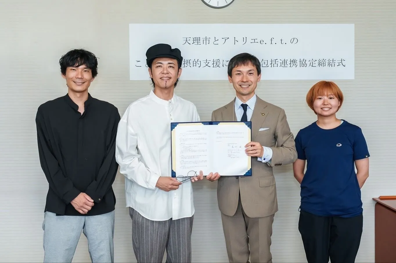 4人の人物が写真に写っています。右から2人目の男性が書類を持っており、他の3人がその周りに立っています。背景には「天理市とアトリエe.f.t. の摂的支援に包括連携協定締結式」と書かれた看板があります。