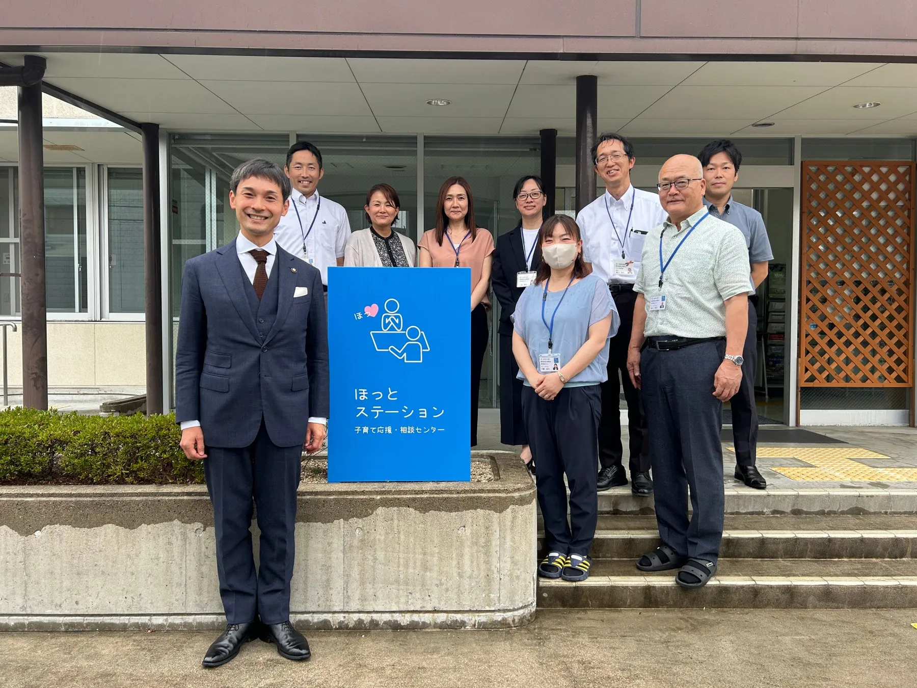 「ほっとステーション 子育て応援・相談センター」と書かれた青い看板の前で記念写真に写る人々。人々は笑顔でカメラを見ています。