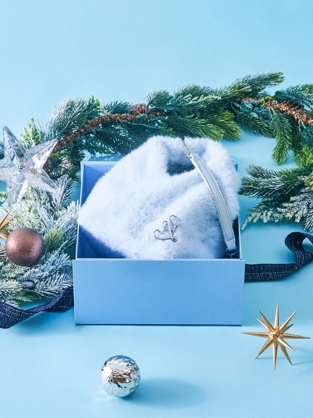 青い箱に入った、水色の毛皮のバッグが写った画像です。バッグの取っ手は白く、ブランドロゴがついています。クリスマスをテーマにした飾り付けが施されており、背景には松の枝、星のオーナメント、銀色のボールなどが配置されています。