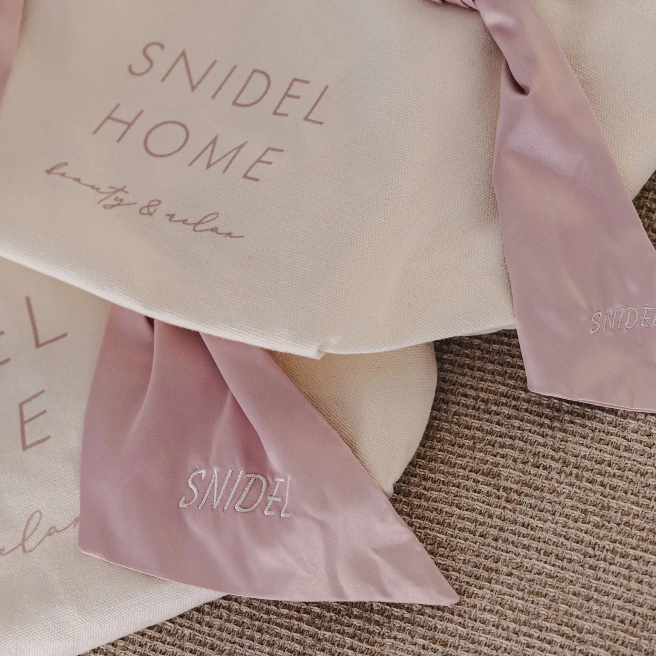 ピンク色のリボンと、ブランドロゴが印字されたバッグ、クッションが写った写真。リボンには「SNIDEL」の文字が刺繍され、背景にはざっくりとした質感の布地が見える。