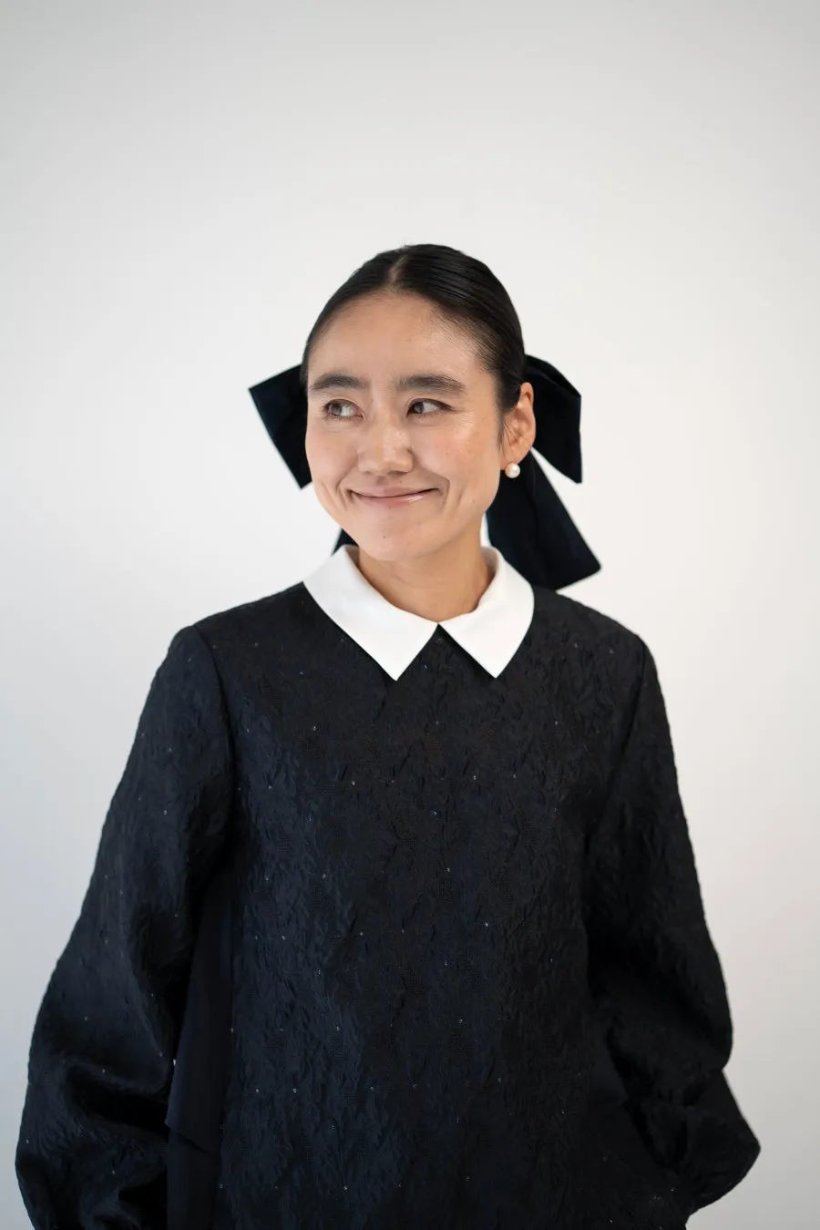 黒いドレスを着て微笑む女性のポートレート。彼女は白い襟と黒いリボンをつけており、背景は白いです。