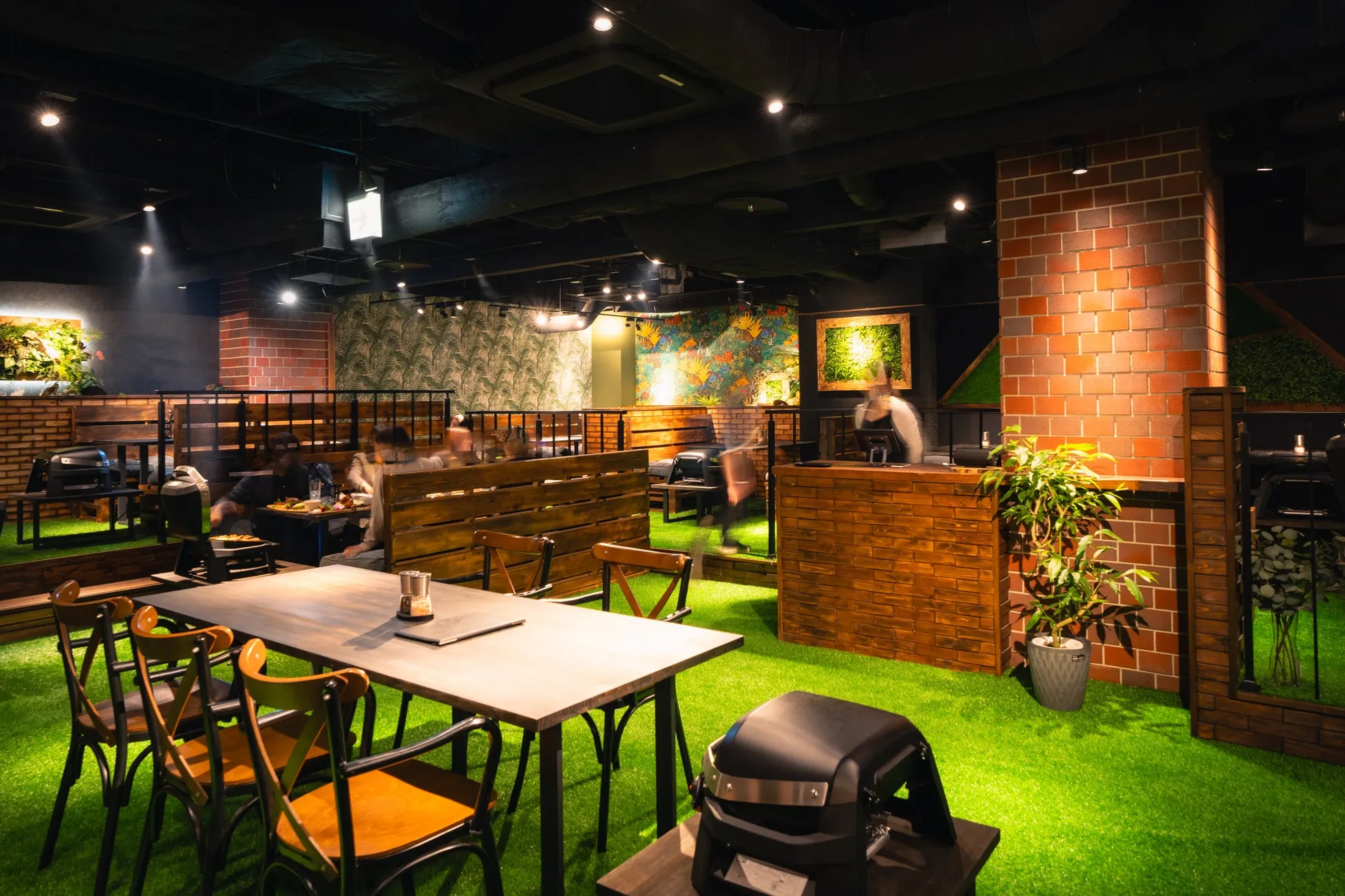 人工芝が敷かれたレストランの内部。レンガや木の壁、緑の装飾が施され、テーブルと椅子が配置されている。人々が食事を楽しんでいる様子が写っている。