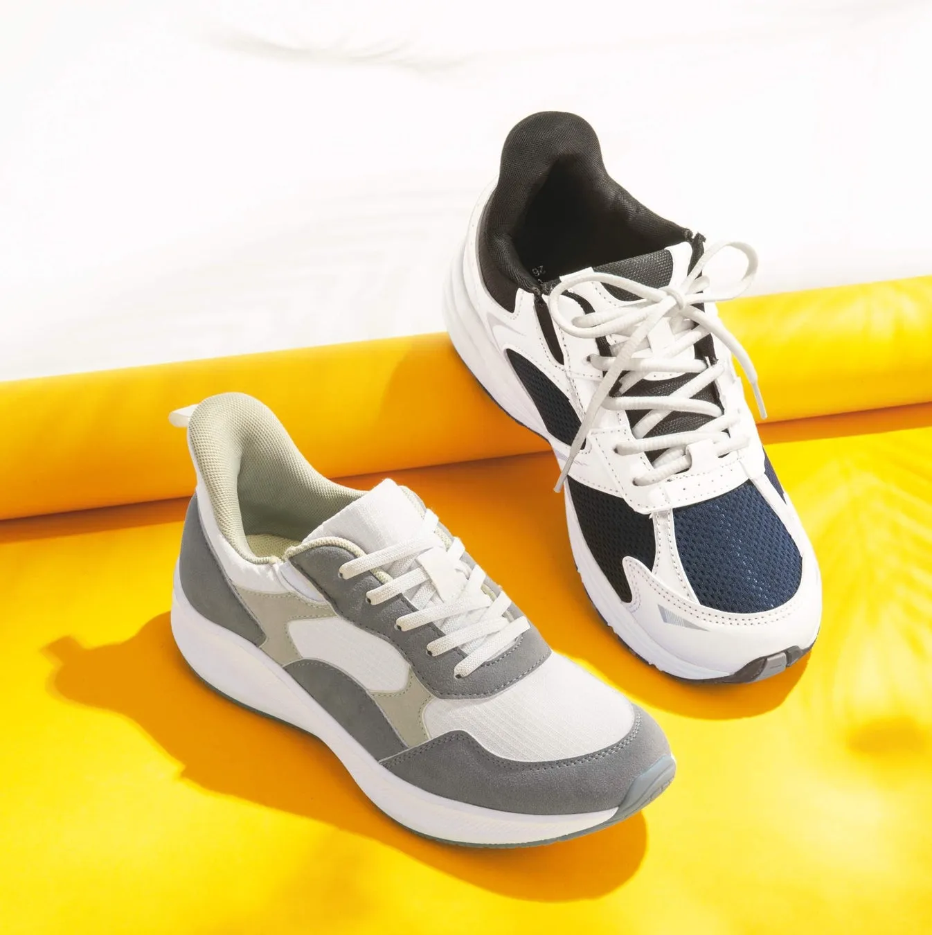 黄色い背景に2つのスニーカーが写っています。1つは白とグレー、もう1つは白とネイビーです。