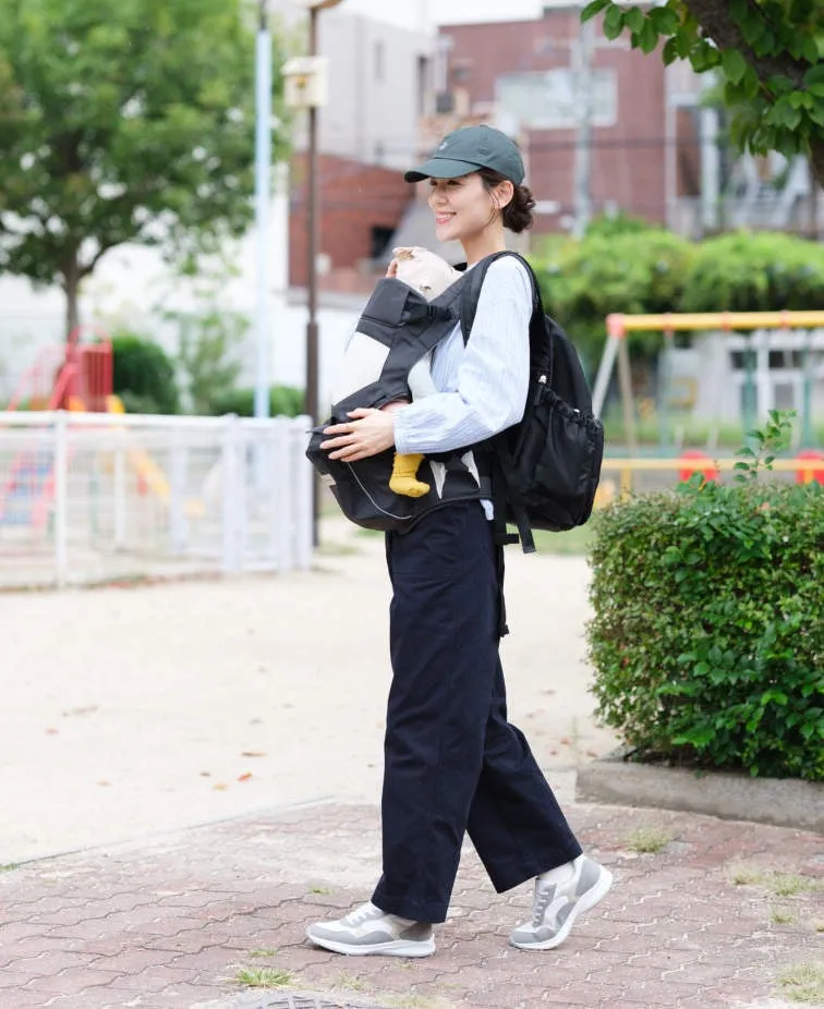 公園で赤ちゃんを抱っこ紐で抱っこしている女性の画像です。女性はキャップをかぶり、リュックを背負い、笑顔で歩いています。背景には遊具があり、公園の様子がわかります。