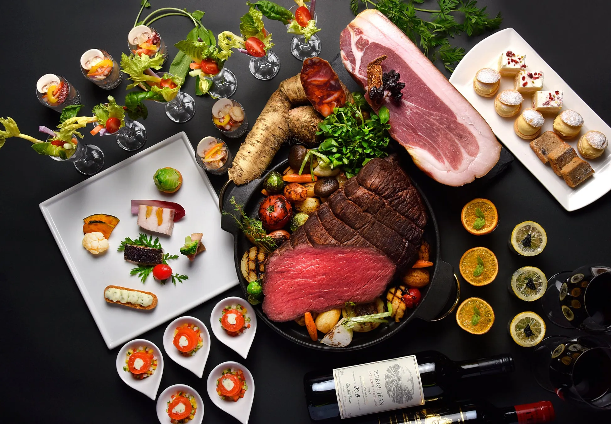 様々な料理が並んだテーブルの俯瞰写真。ローストビーフ、生ハム、前菜、デザート、ワイン、グラスなどが配置され、高級感のある食事の様子が写し出されています。