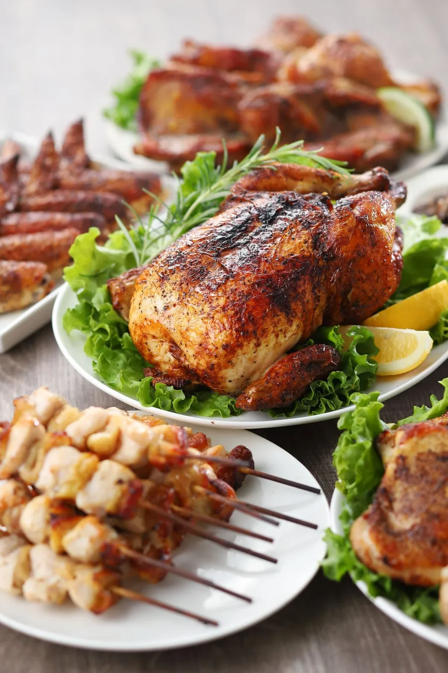 テーブルの上に、ローストチキン、チキンウイング、鶏肉の串焼きなど、さまざまな種類の鶏肉料理が並んだ写真です。各料理は、レタスの上に盛り付けられ、レモンやハーブなどの飾りで彩られています。