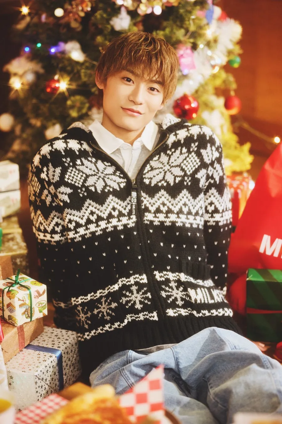 クリスマスツリーの前に座っている男性。黒と白のセーター、プレゼントの山の画像