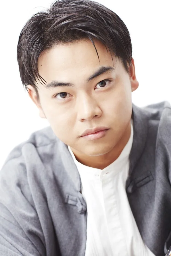 正面を向いた若い男性のポートレート。彼はグレーのジャケットと白いシャツを着ており、カメラ目線で少し真面目な表情をしている。