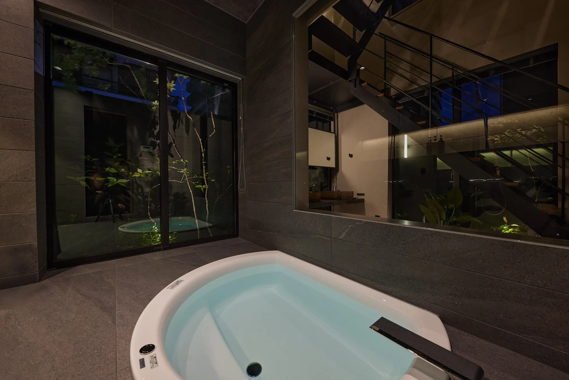 浴槽と窓が特徴的なモダンなバスルームの写真。窓からは庭が見え、階段と鏡が奥に見える。