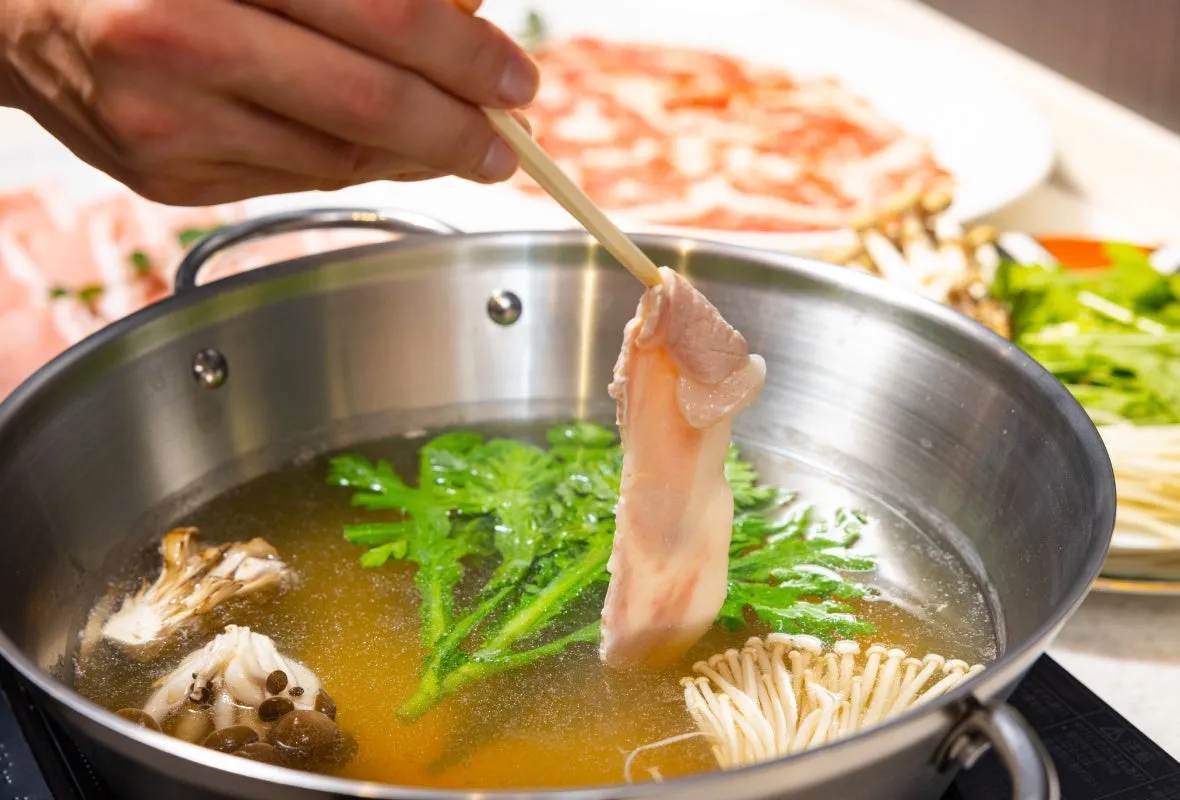 鍋料理の様子。箸で豚肉を掴み、出汁の中にくぐらせている。鍋の中には野菜やキノコが入っている。
