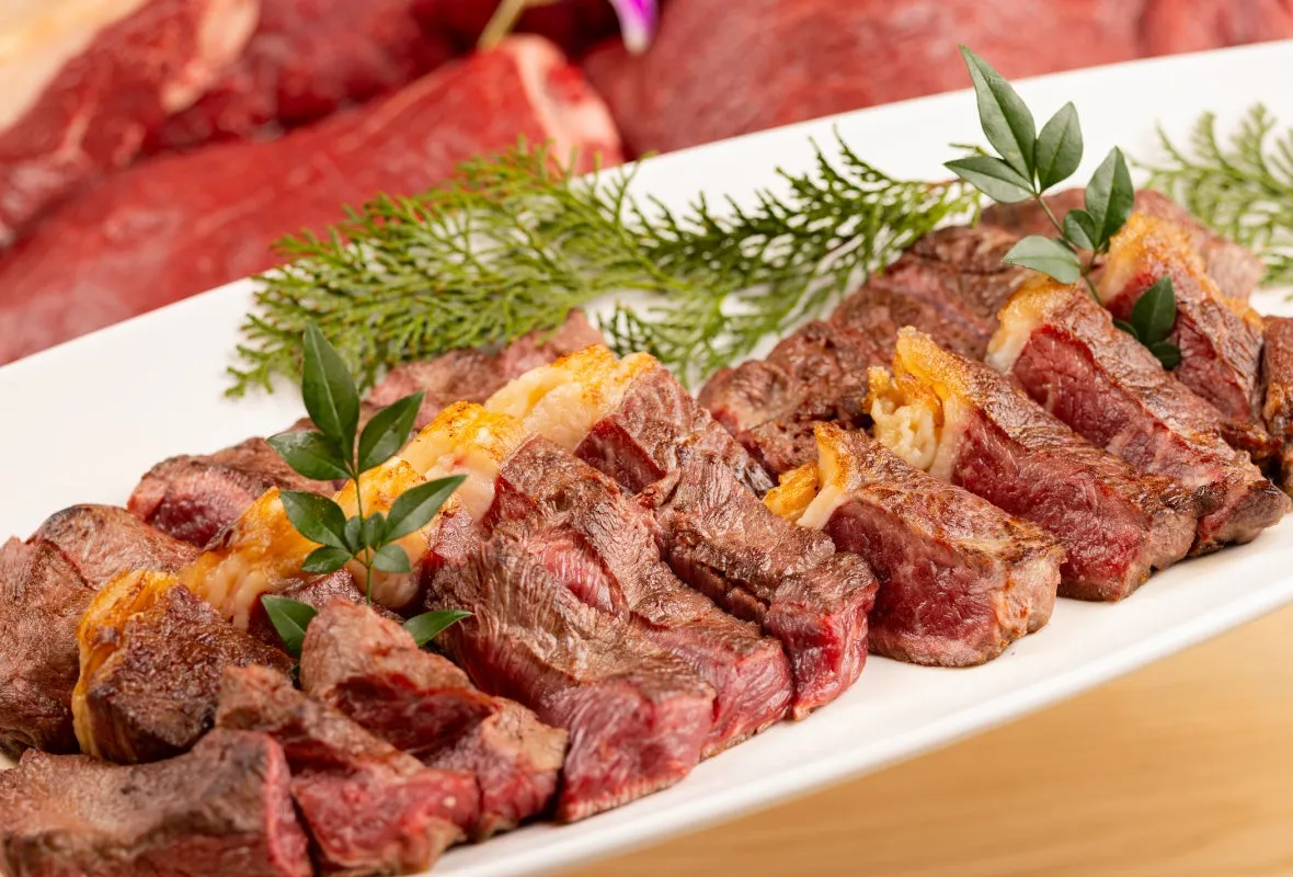 お皿に盛られたステーキのクローズアップ写真。ステーキはカットされ、いくつかの緑の葉で飾られています。背景には、未調理の肉の塊があります。