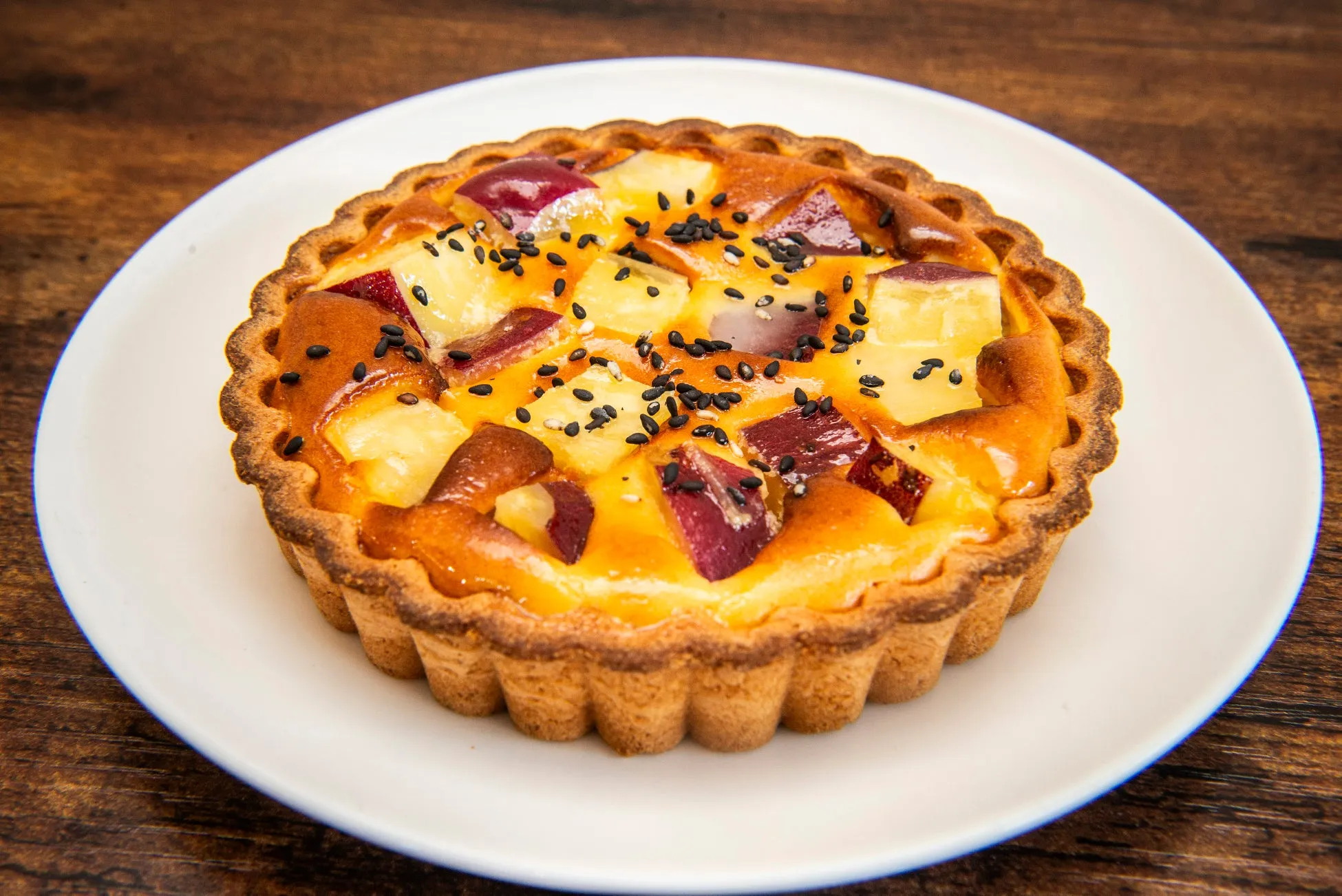 お皿に載った、さつまいもとパイナップルのタルトのクローズアップ写真。タルトは、さつまいもとパイナップルの塊、黒いゴマがトッピングされています。背景は木製の表面です。