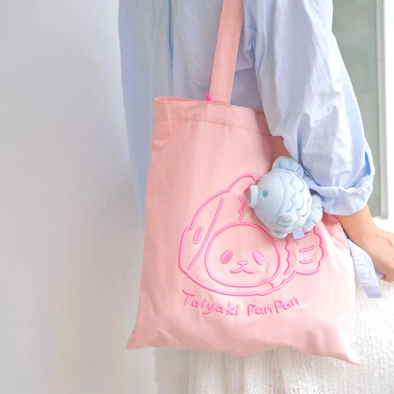 ピンク色のトートバッグを肩にかける女性。バッグにはTaiyaki Pan Panのキャラクターが刺繍されており、青い魚のぬいぐるみも付いている。