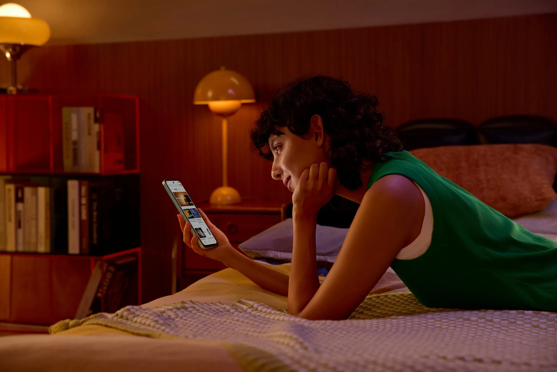 ベッドに横たわる女性がスマートフォンを見ています。スマートフォンにはYouTubeの画面が表示されています。部屋は暖色系の照明で照らされています。