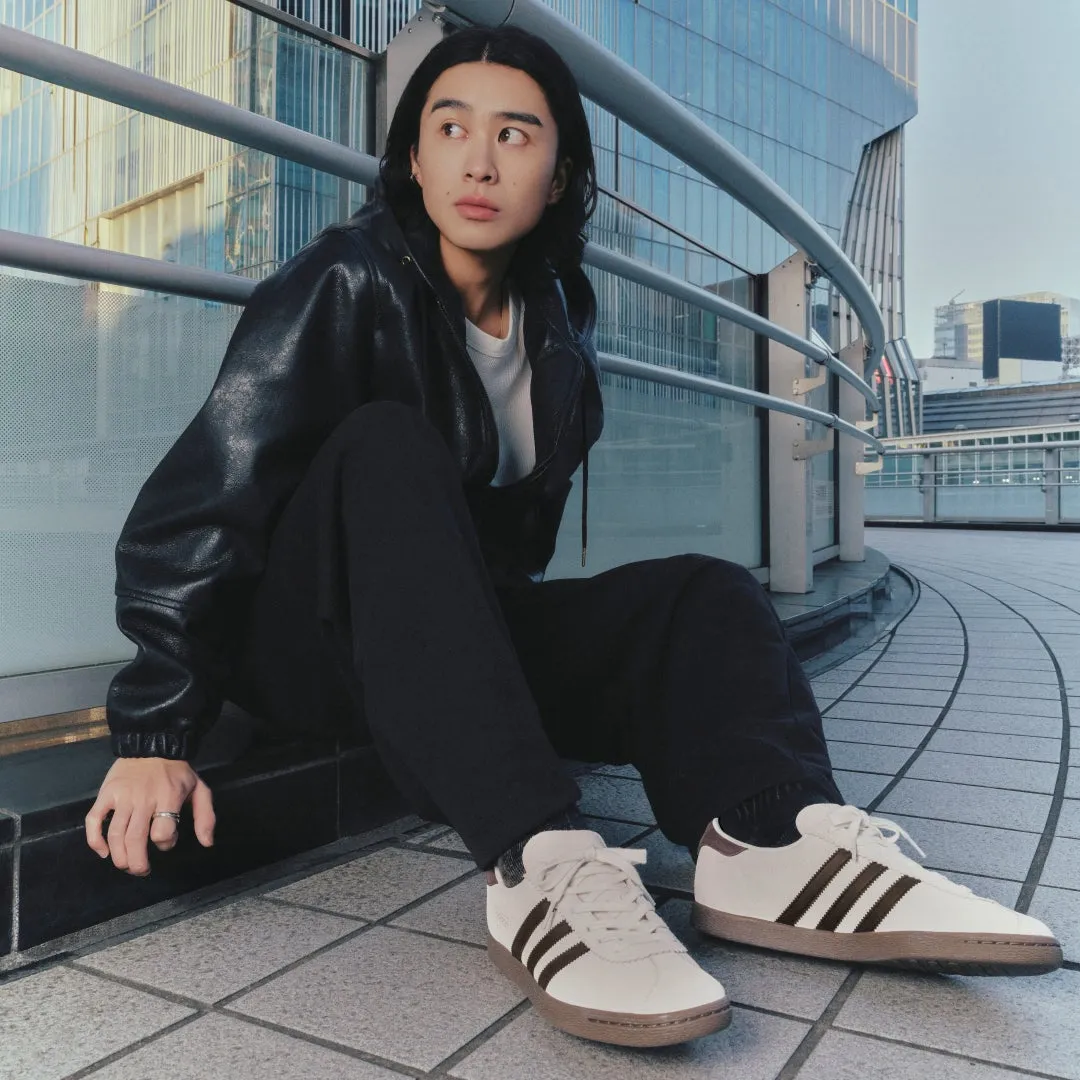 男性が屋外で座って見上げる様子。彼は黒いジャケットとズボンを着ており、白いスニーカーを履いている。背景にはガラス張りの建物が写っている。