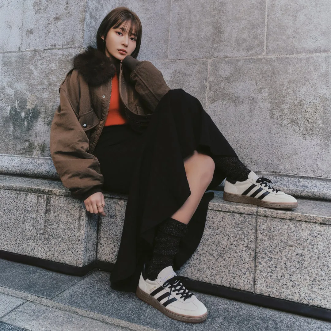 女性が座ってポーズをとっているファッションフォト。彼女は茶色のジャケットと黒いスカートを着ており、アディダスのスニーカーを履いている。背景はコンクリートの壁。