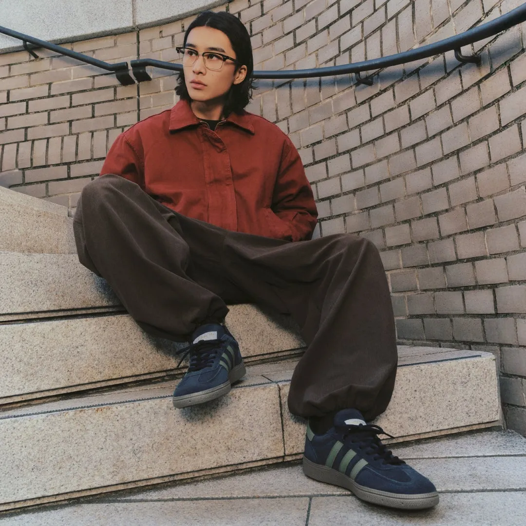 階段に座っている男性。赤いジャケットと茶色のパンツを着ており、メガネをかけている。足元にはアディダスのスニーカー。